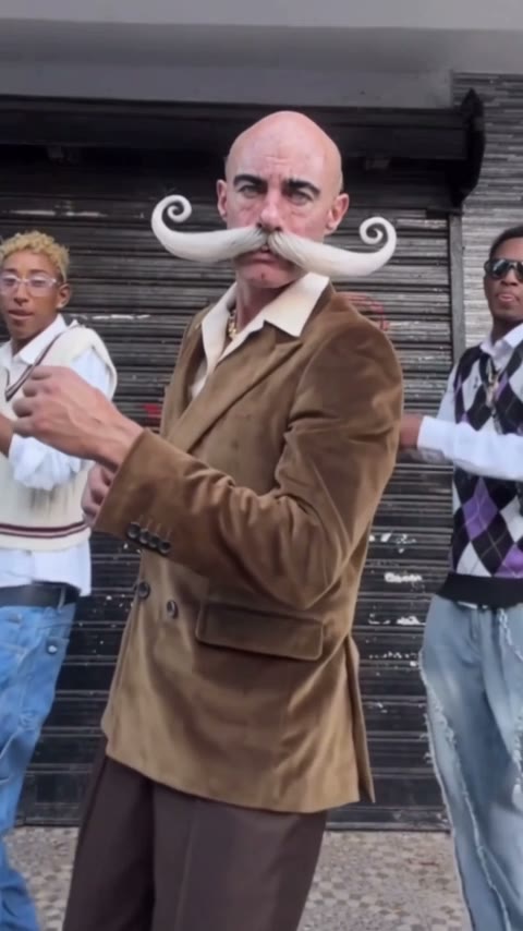 A Dame Hombro AI video made from one photo: a bald man with a curled white moustache in a brown velvet suit dancing between two dancers